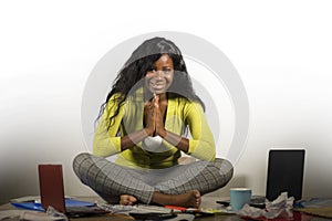 Young happy and attractive African American business woman doing yoga sitting at office messy desk full of paperwork smiling relax