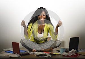 Young happy and attractive African American business woman doing yoga sitting at office messy desk full of paperwork smiling relax