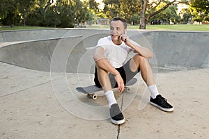 Young happy and attractive American man 30s sitting on skate board after sport boarding training session talking on mobile phone
