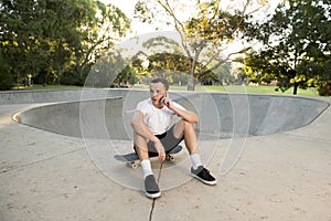 Young happy and attractive American man 30s sitting on skate board after sport boarding training session talking on mobile phone