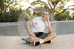 Young happy and attractive American man 30s sitting on skate board after sport boarding training session talking on mobile phone