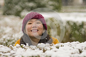 Young happy and attractive Asian Chinese woman in Winter jacket and beanie enjoying snowfall at city park playing cheerful