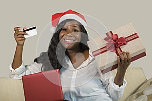 Young happy and attractive black African American woman in Santa Klaus hat holding credit card and Christmas present box doing onl