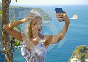 Young happy and attractive blond woman taking selfie portrait with mobile phone at beautiful tropical paradise view of ocean rock