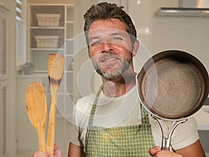 Young happy and attractive home cook man in apron smiling cheerful and charming holding pan and spoon enjoying domestic work and