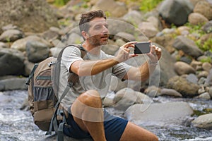 Young happy and attractive man taking landscape photo with mobile phone holding backpack hiking in river at forest enjoying nature