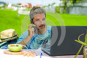 Young happy and attractive man working outdoors with laptop computer and mobile phone as internet travel blogger or digital nomad
