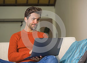 Young happy and attractive man working relaxed with laptop computer at modern apartment living room sitting at sofa couch typing a