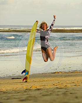 Young happy and attractive surfer girl jumping high in the air holding surf board before surfing at beautiful tropical beach enjoy