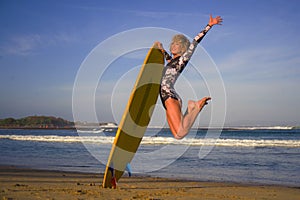 Young happy and attractive surfer girl jumping high in the air holding surf board before surfing at beautiful tropical beach enjoy