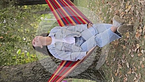 Young happy attractive woman enjoying relaxation sitting in fabric hammock chair in park. Vertical video