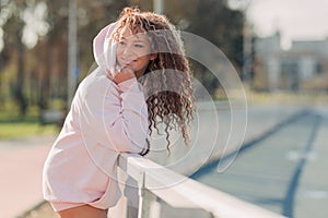 Young happy attractive woman posing on a fence near a running track