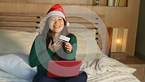 Young happy beautiful Asian American girl on bed in Santa Christmas hat using credit card and laptop computer for online shopping