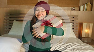 Young happy beautiful Asian American girl in winter hat holding Christmas giftbox with ribbon smiling excited and cheerful receivi