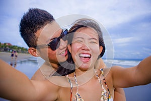 young happy and beautiful Asian Chinese couple taking selfie photo with mobile phone camera smiling joyful having fun on the beach