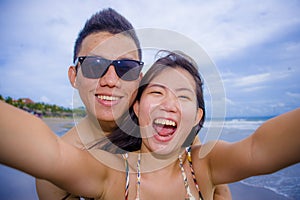 young happy and beautiful Asian Chinese couple taking selfie photo with mobile phone camera smiling joyful having fun on the beach