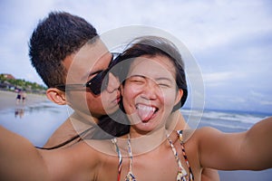 young happy and beautiful Asian Chinese couple taking selfie photo with mobile phone camera smiling joyful having fun on the beach