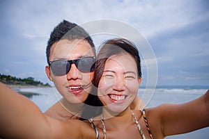 young happy and beautiful Asian Chinese couple taking selfie photo with mobile phone camera smiling joyful having fun on the beach