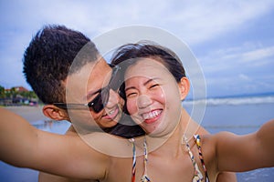 young happy and beautiful Asian Chinese couple taking selfie photo with mobile phone camera smiling joyful having fun on the beach