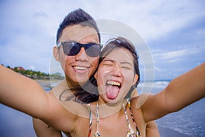 young happy and beautiful Asian Chinese couple taking selfie photo with mobile phone camera smiling joyful having fun on the beach