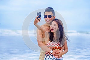 young happy and beautiful Asian Chinese couple taking selfie photo with mobile phone camera smiling joyful having fun on the beach