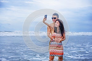 young happy and beautiful Asian Chinese couple taking selfie photo with mobile phone camera smiling joyful having fun on the beach