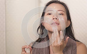 Young happy and beautiful Asian Chinese woman applying moisturizer facial cream and beauty treatment at home bathroom in morning
