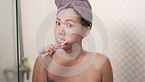Young happy and beautiful Asian Chinese woman brushing her teeth wearing head towel wrap at bathroom mirror