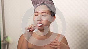 Young happy and beautiful Asian Chinese woman brushing her teeth wearing head towel wrap at bathroom mirror