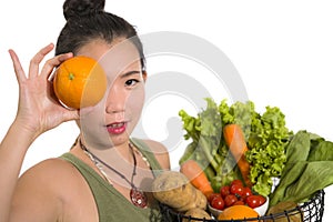 Young happy and beautiful Asian Chinese woman holding basket full of vegetables smiling cheerful holding orange playful in healthy