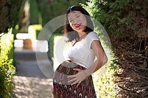 Young happy and beautiful Asian Chinese woman posing outdoors happy and cheerful at city park pregnant showing her belly proud