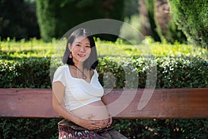 Young happy and beautiful Asian Chinese woman pregnant showing her belly proud and cheerful smiling at city park bench happy in