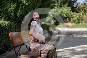 Young happy and beautiful Asian Chinese woman pregnant showing her belly proud and cheerful smiling at city park bench happy in