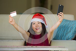 Young happy and beautiful Asian Chinese woman in Santa Claus hat holding credit card using mobile phone for Christmas online shopp