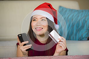 Young happy and beautiful Asian Chinese woman in Santa Claus hat holding credit card using mobile phone for Christmas online shopp