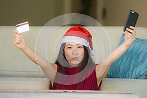 Young happy and beautiful Asian Chinese woman in Santa Claus hat holding credit card using mobile phone for Christmas online shopp