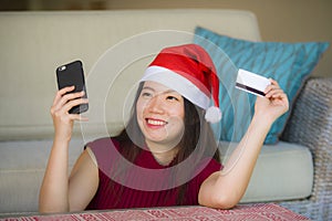 Young happy and beautiful Asian Chinese woman in Santa Claus hat holding credit card using mobile phone for Christmas online shopp