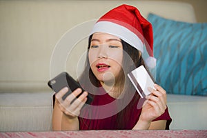 Young happy and beautiful Asian Chinese woman in Santa Claus hat holding credit card using mobile phone for Christmas online shopp