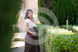 Young happy and beautiful Asian Japanese woman posing outdoors happy and cheerful at city park pregnant showing her belly proud