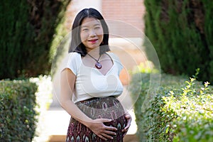 Young happy and beautiful Asian Japanese woman posing outdoors happy and cheerful at city park pregnant showing her belly proud