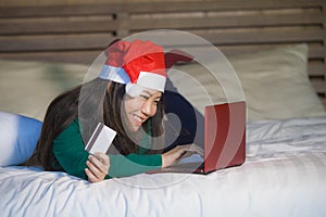 Young happy and beautiful Asian Korean girl on bed in Santa Christmas hat using credit card and laptop computer for online shoppin