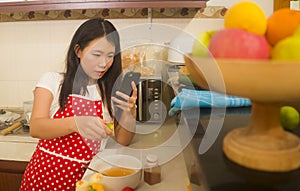 Young happy and beautiful Asian Korean home cook woman in red apron reading healthy recipe in mobile phone cooking cheerful
