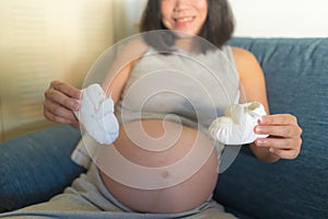 Young happy and beautiful Asian Korean woman pregnant on couch enjoying playful with little baby booties smiling relaxed at home