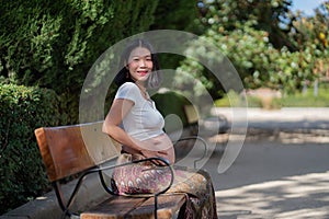 Young happy and beautiful Asian Korean woman pregnant showing her belly proud and cheerful smiling at city park bench happy in