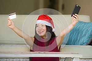 Young happy and beautiful Asian Korean woman in Santa Claus hat holding credit card using mobile phone for Christmas online shoppi