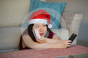 Young happy and beautiful Asian Korean woman in Santa Claus hat holding credit card using mobile phone for Christmas online shoppi