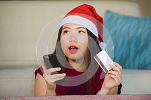 Young happy and beautiful Asian Korean woman in Santa Claus hat holding credit card using mobile phone for Christmas online shoppi