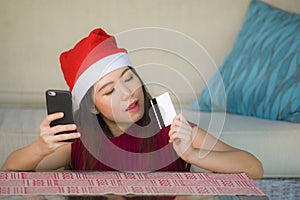 Young happy and beautiful Asian Korean woman in Santa Claus hat holding credit card using mobile phone for Christmas online shoppi