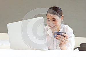 Young happy beautiful Asian woman using laptop computer and holding credit card while lying on the bed, businesswoman working