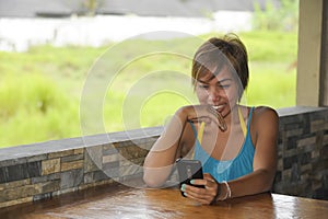 Young happy beautiful and attractive Asian woman sitting in coffee shop using internet app on mobile phone laughing excited outdoo